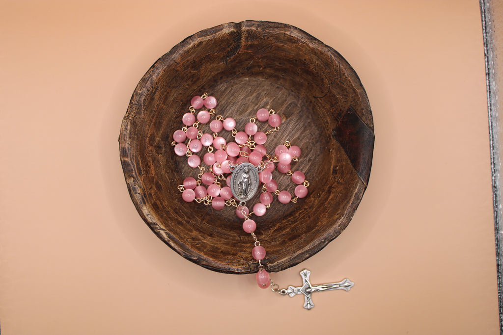 Pink rosary with a silver cross on a wooden bowl against a beige background